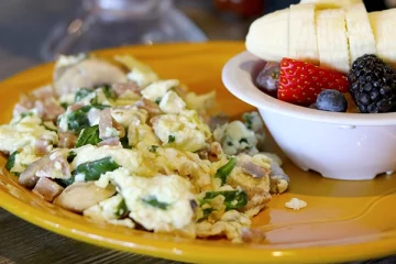 A yellow plate with scrambled eggs mixed with spinach and mushrooms next to a bowl of sliced bananas, strawberries, blueberries, and blackberries.